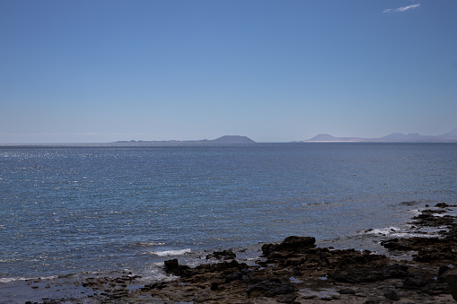Rocky coast at the south of Lanzarote. Calm water of Atlantic ocean. Fuerteventura island on the horizon. Blue sky with white clouds. Playa Blanca, Lanzarote, Canary Islands, Spain.