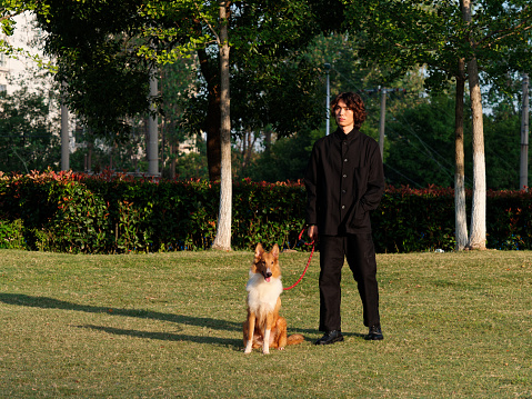 Portrait of a handsome Chinese young man in black with his rough collie dog on green grass field in sunny day, male fashion, cool Asian young man lifestyle, harmony man and pet.