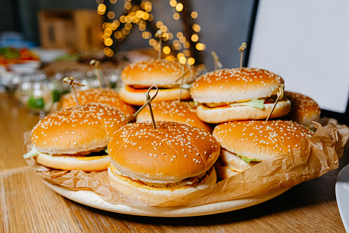 A platter of delicious burgers topped with sesame seeds, presented on a table with warm, festive bokeh lights in the background