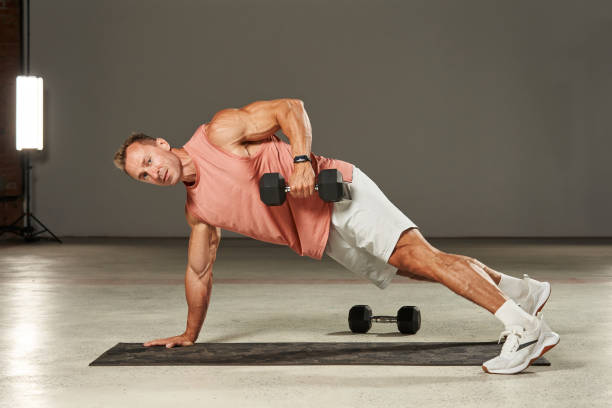 istock Caucasian athlete stands in a plank position on the mat on one arm and pulls a large dumbbell to his chest with the other hand. fitness. aerobics. exercises on the mat. physical health 1904450479
