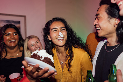 A group of young people enjoying a party at a house in North Shields, North East England. A woman is celebrating her birthday and a friend has just cake smashed her. She has cream on her nose.