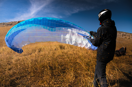 A paraglider lands or rises on the yellow grass on a sunny day. A man after or before a paragliding flight. Glider with parachute, wide angle rear view