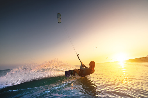 A girl kiter rides against a beautiful background of splashes and a colorful sunset of the sea. Woman kitesurfing on the water with a fantastic view in the background