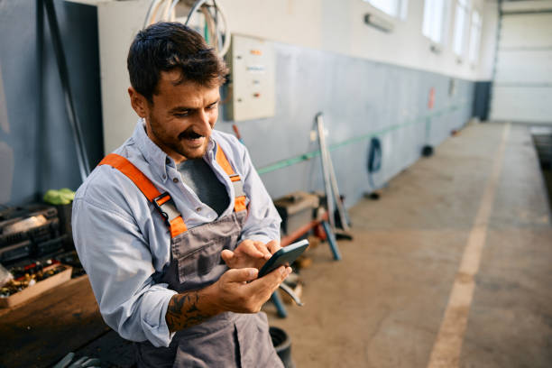 Happy mechanic texting on cell phone while working in a garage. Happy mechanic using smart phone while working in a repair shop. Copy space. working class stock pictures, royalty-free photos & images