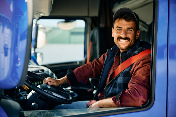 Happy professional truck driver driving his truck and looking at camera. Happy truck driver looking through side window while driving his truck. truck driver stock pictures, royalty-free photos & images