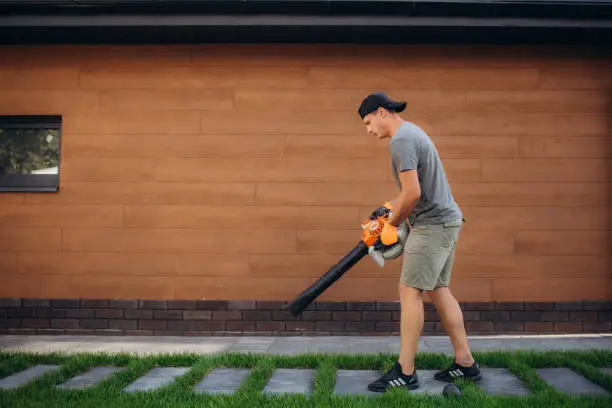 man uses a blower, a vacuum cleaner works in an autumn garden, blowing off fallen leaves from a garden path. High quality photo