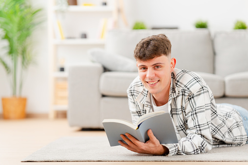 Portrait of happy young caucasian student looking at camera. Teenager boy reading a book lying on the carpet