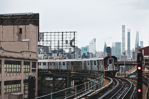 New York City subway train is approaching an elevated subway station in Queens. Raining day in New York. Manhattan skyline in the background.