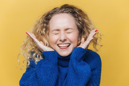 Joyful woman in a blue sweater laughing with eyes closed, her hands by her face, against a yellow background