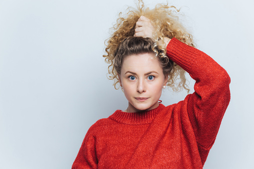 Curious woman with voluminous curly hair in a vibrant red sweater, hand in hair, looking thoughtfully aside