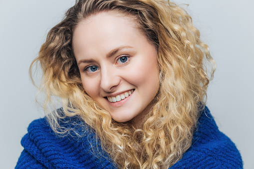 Cheerful young woman with vibrant curly blonde hair, beaming smile, wrapped in a cozy blue knit sweater.