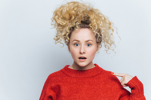 Surprised young woman with wild curly hair, pointing at herself in disbelief, wearing a vivid red sweater