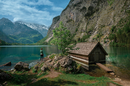 Woman standing at boathouse with mountain cliffs on lake Obersee at Berchtesgaden Bavaria, clouds in blue sky, turquoise calm water, Berchtesgaden Bavaria