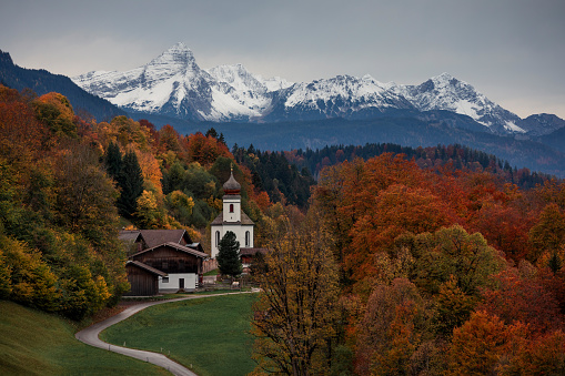 Bavarian Alps with church of Wamberg in Garmisch-Partenkirchen during autumn, snow-covered mountains in the background, dramatic cloudy sky, colored leaves and trees and road in foreground, Bavaria Germany