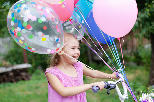 Little girl gets bicycle with balloons. Kid with gift or present. Child is smiling, having fun. Celebration of Happy Birthday party outside in summer backyard. Lifestyle moment, candid real emotions.