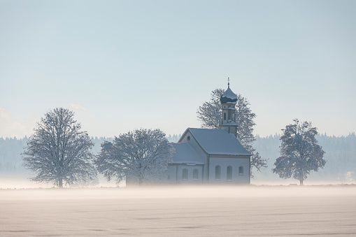 Bavarian church of Raisting with trees and snow and mist during winter, snow field in the foreground, blue sky day, Bavaria Germany