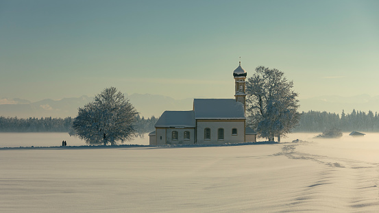 Bavarian church of Raisting with trees and snow and mist during winter, snow field in the foreground, blue sky day, Bavaria Germany