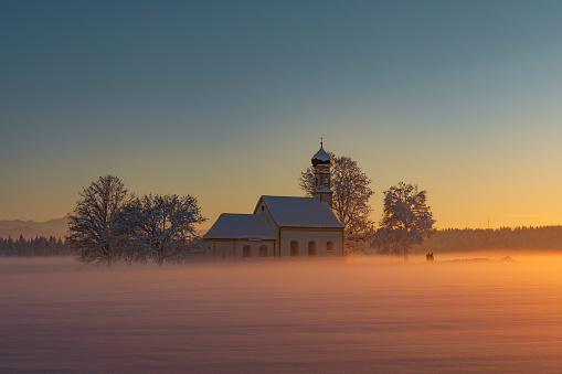 Bavarian church of Raisting with trees and snow and mist during winter and sunset, snow field in the foreground, blue sky day, Bavaria Germany
