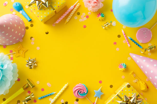 Lovely birthday display inspiration. Top-view image capturing table with joyful decor, merengue sweets, gifts, hats, candles, balloons, confetti, serpentine on yellow backdrop. Text-friendly space