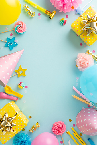 Joyous birthday party concept. Overhead vertical shot of cheerful table arrangement featuring presents, birthday hats, lollipops, balloons, confetti on pastel blue background. Perfect for text or ad