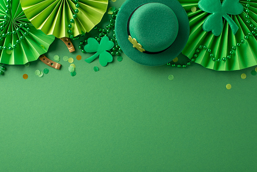 Top-view of festive Saint Patrick's Day setup: leprechaun's hat, fortunate horseshoe, cheering fans, clovers, confetti, and beaded necklaces on vibrant green backdrop. Ad-ready with space for message