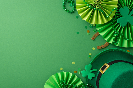 Captivating Saint Patrick's Day top view composition: leprechaun's hat, lucky horseshoe, festive fans, trefoils, confetti, beads necklace set on lively green backdrop, offering space for text or promo