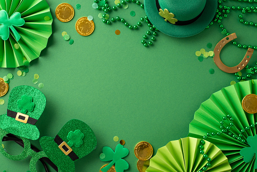 Festive St Patrick's top view scene: leprechaun's hat, whimsical glasses, lucky horseshoe, gold coins, fans, clovers, confetti, beads necklace arranged on green surface, leaving space for your message