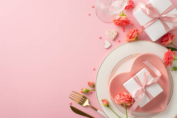 istock Anniversary Elegance: top view sophisticated table setting with heart-shaped plate, cutlery, wineglass, roses, gift boxes on pink background. Ideal ambiance for celebrating love. Text space available 1904963144
