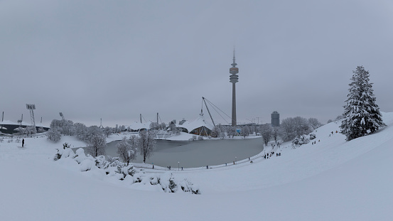 Panorama of Munich Olympiapark covered in snow during winter, snowfall and clouds in the sky, TV tower, icy Olympia lake and Olympia stadion, people skiing on the hills, 02 Feb 2023