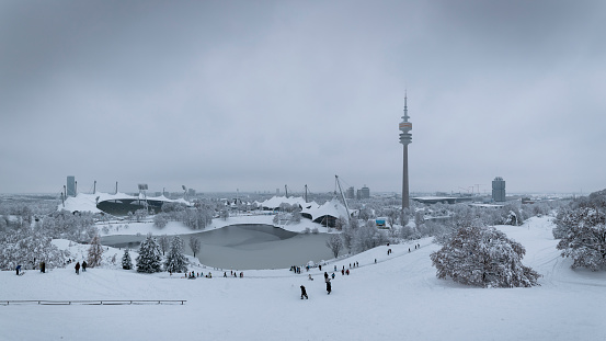 Panorama of Munich Olympiapark covered in snow during winter, snowfall and clouds in the sky, TV tower, icy Olympia lake and Olympia stadion, people skiing on the hills, 02 Feb 2023