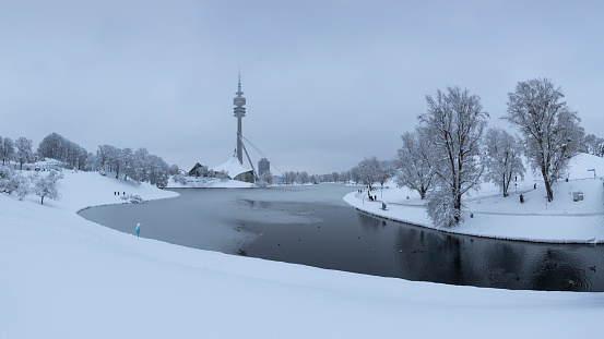Panorama of Munich Olympiapark covered in snow during winter, snowfall and clouds in the sky, TV tower and icy Olympia lake, 02 Feb 2023