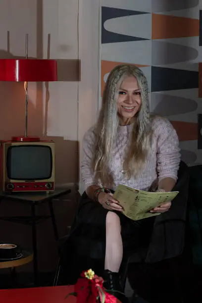 Platinum blonde girl, 25-30 years old, wearing a black leather skirt and pink sweater, sitting reading the menu of a 70s-style restaurant. With a red lamp and an antique television behind her.