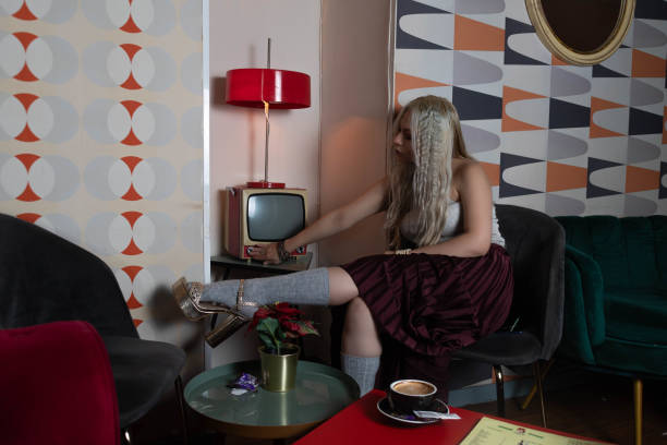 プラチナブロンドの女の子、25〜30歳、白いタンクトップと赤いスカート、灰色のストッキングを着て、アンティークの茶色の椅子に座っています。古いテレビをつける。テーブルの上にコー - 25 30 years audio ストックフォトと画像