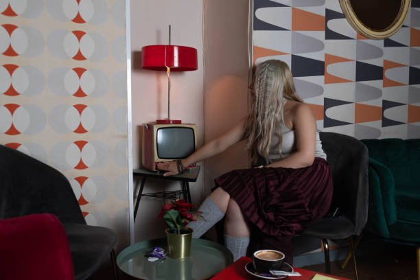 platinum blonde girl, 25 to 30 years old, dressed in a white tank top and a red skirt, gray stockings, sitting on an antique brown chair. turning on an old television. with a cup of coffee on the table. side view.. in a 70s style restaurant. - side table table antique classic fotografías e imágenes de stock