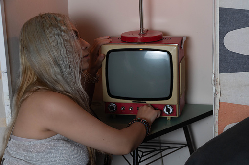 Platinum blonde girl, 25 to 30 years old, wearing a white tank top. Turning on an old television. Side view.. In a 70s style restaurant.
