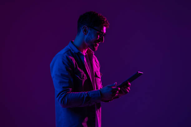 istock Young man with modern digital device isolated over gradient blue studio background in pink neon light. 1905031304