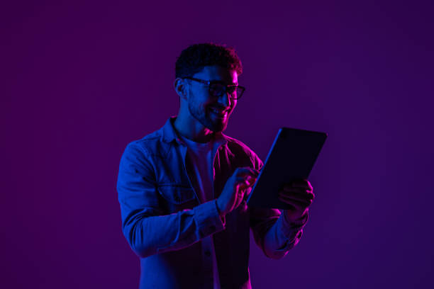 istock Young man with modern digital device isolated over gradient blue studio background in pink neon light. 1905036478