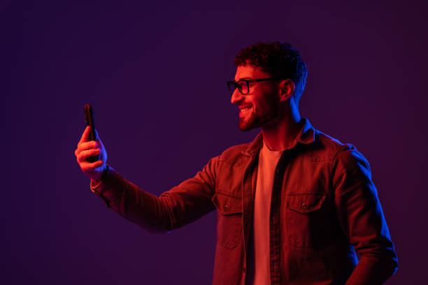 istock Concentrated male browsing mobile phone while standing on black background with glowing blue and pink neon lights 1905036994
