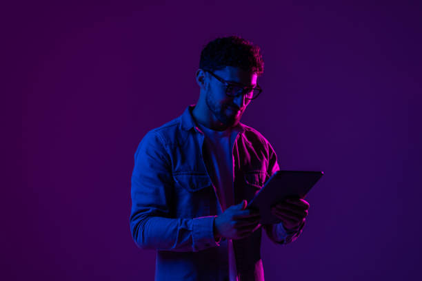 istock Young man with modern digital device isolated over gradient blue studio background in pink neon light. 1905037505