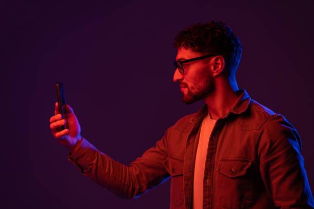 istock Concentrated male browsing mobile phone while standing on black background with glowing blue and pink neon lights 1905038551