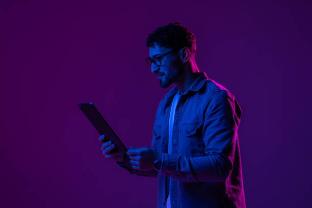 istock Young man with modern digital device isolated over gradient blue studio background in pink neon light. 1905040161