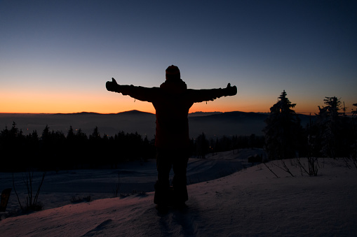 Silhouette of alone tourist standing on snowy mountain top in winner pose with raised hands enjoying beautiful sunset. Adventure, outdoors activities, healthy lifestyle.