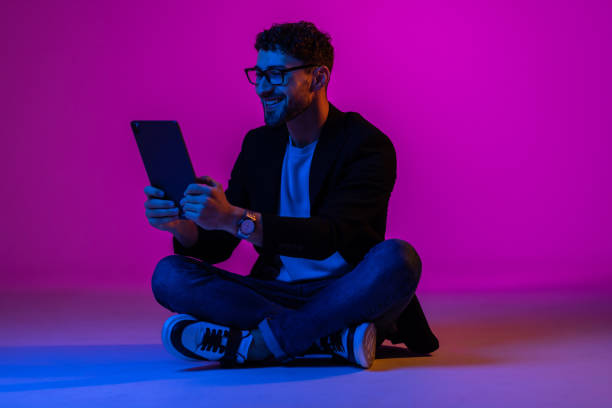 istock Young man wearing business suit and eyeglasses using digital tablet on gradient blue purple background in neon light. 1905205468