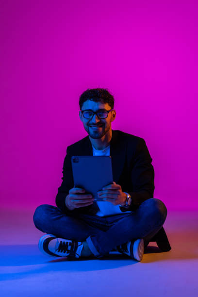 istock Young man wearing business suit and eyeglasses using digital tablet on gradient blue purple background in neon light. 1905212254