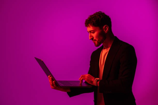istock Portrait of man with portable computer in hand, looking at display on neon light background. 1905213291