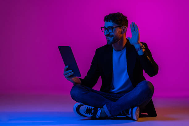 istock Young man wearing business suit and eyeglasses using digital tablet on gradient blue purple background in neon light. 1905221165
