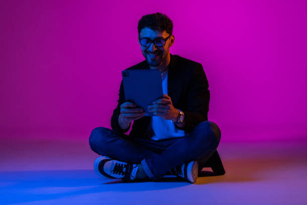 istock Young man wearing business suit and eyeglasses using digital tablet on gradient blue purple background in neon light. 1905221681