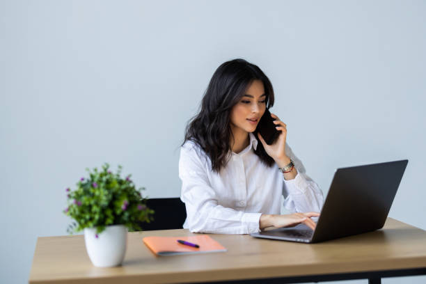 istock Young beautiful woman in glasses talking on mobile phone and using laptop with smile while sitting at her working place 1905255485