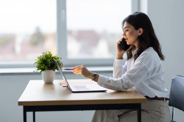 istock Young beautiful woman in glasses talking on mobile phone and using laptop with smile while sitting at her working place 1905263694
