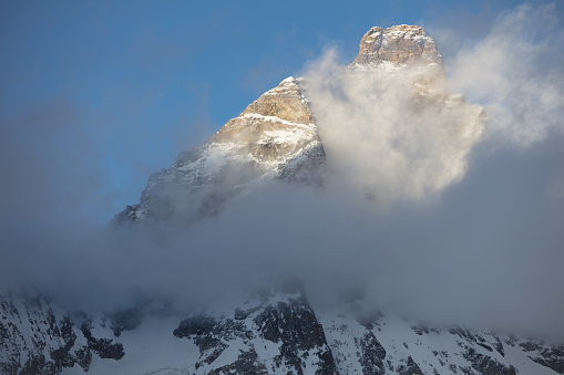 It is a late winter afternoon, the light is dim, and the mountain peaks are shrouded in clouds. The Mattherhorn's summit, resembling a granite pyramid, still lit by the setting sun, is clearing from the clouds.